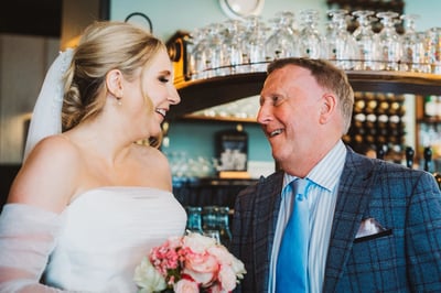 A joyful bride in a white strapless dress and pink bouquet stands in front of a man in a blue plaid suit, both smiling and engaged in a conversation. The man holds a bouquet of pink and white flowers, and the background features a bar with wine glasses and a clock. The scene captures a special moment between the bride and the man, set against a warm and inviting atmosphere.
