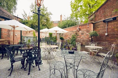 The image shows a serene courtyard with black metal tables and chairs, white umbrellas, and a brick wall with a chimney. The ground is paved with cobblestones, and potted plants add greenery. The scene is peaceful and inviting, with a sense of tranquility.