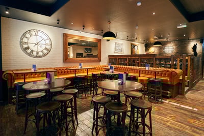 The image shows a modern bar with a wooden floor, white brick wall, and a large clock. The bar has six wooden stools and a wooden bar, with blue napkins on the stools. The wall has a mirror and a framed picture, and the ceiling has black pendant lights. The bar is illuminated by a black lamp, and the overall atmosphere is warm and inviting.