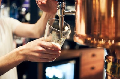 A person in a white shirt is pouring a frothy, frothy white liquid into a clear glass with a silver spout, using a copper-colored glass distillation machine. The background is blurred, suggesting a bar or restaurant setting. The image captures a moment of skill and precision in the craft of distilling.