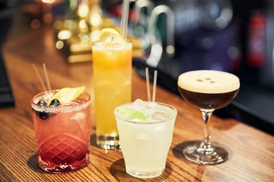 Three drinks are displayed on a wooden bar, each with a unique color and garnish. The first drink is a vibrant yellow, with a lime wedge and a slice of lemon. The second drink is a dark red, topped with a slice of orange and a lime wedge. The third drink is a pale yellow, with a slice of lemon and a lime wedge. The background is blurred, suggesting a busy bar. The arrangement creates a visually appealing display.