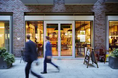 A brick building with large windows and a glass door is the focal point of a bustling street scene. Two individuals walk past the entrance, while a wooden signboard and potted plant add to the urban atmosphere. The scene is bathed in warm, inviting light, creating a lively and inviting atmosphere.