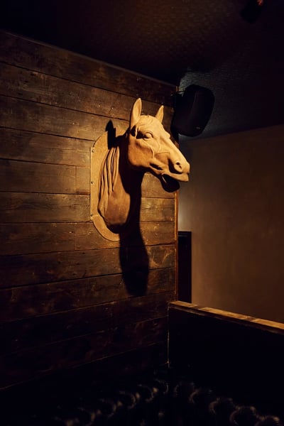 A wooden wall with a horse head sculpture is illuminated by a hanging lamp, casting a warm glow on the wall and creating a cozy atmosphere. A black leather couch is positioned to the right, inviting relaxation. The composition is balanced and harmonious, with the horse head sculpture as the focal point.