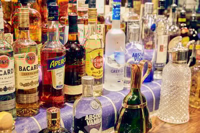 The image features a diverse collection of 20+ liquor bottles, each with unique labels and colors, arranged on a blue and gold tablecloth. The bottles are arranged in a semi-circle, with some overlapping each other. The background is blurred, focusing on the bottles and their arrangement. The image is taken from a high angle, showcasing the bottles' design and color. The scene evokes a sense of abundance and variety, typical of a bar or restaurant setting.