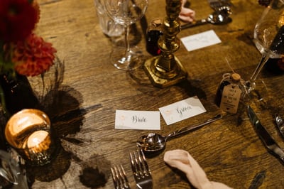 The image shows a beautifully set table for a formal dining event, with a wooden table, silverware, and candle. The table is adorned with a gold candle holder, a candle, and a vase of red and orange flowers. The arrangement suggests a well-thought-out table setting for a special occasion.