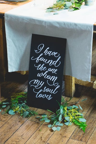 A beautifully set table features a white tablecloth, a black sign with a white cursive message, and a floral arrangement of greenery. The scene is set in a room with wooden flooring, creating a warm and inviting atmosphere.