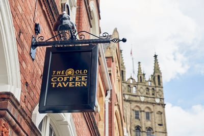 The image features a brick building with a black signboard displaying "THE GROVE CAFE" and "Cafe 100". A black lamp post is attached to the signboard, and a Gothic spire is visible in the background. The scene is set against a clear blue sky with white clouds, creating a serene atmosphere.