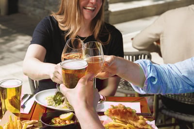 A lively outdoor dining scene features a group of people enjoying a toast, with a woman in a blue shirt and a man in a light blue shirt. They are surrounded by various food items, including a plate of food, a bowl of fries, a glass of beer, and a plate of bread. The table is set with a variety of glasses, including a tall glass of beer and a small glass of wine. The scene is set against a backdrop of a stone wall and a wooden table, creating a warm and inviting atmosphere.
