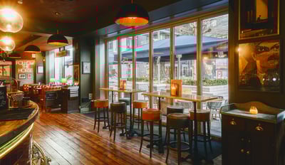 The image shows a cozy, well-lit bar with a wooden counter, four bar stools, and a wooden bar. The bar is adorned with candles and a blue umbrella, and the walls are painted green. Large windows offer a view of a garden, and the bar is illuminated by hanging lamps. The scene is inviting and cozy, with a harmonious blend of colors and textures.