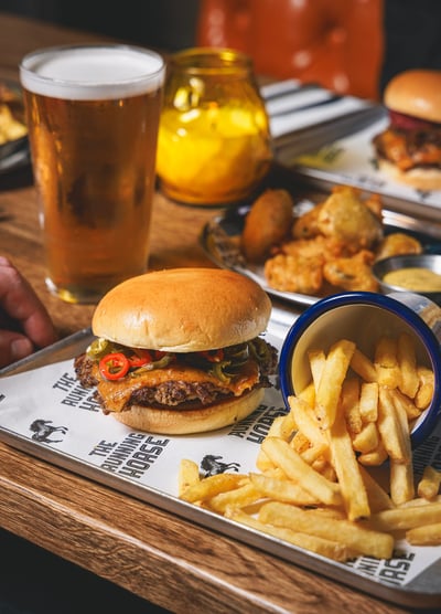 The image features a wooden table with a variety of food items, including a large burger with a bun, meat, and red sauce, a side of fries, a glass of beer, a blue bowl with a pickle, and a tray with additional food items. The scene is set in a restaurant or diner, with a hand visible, suggesting someone is about to enjoy the meal.