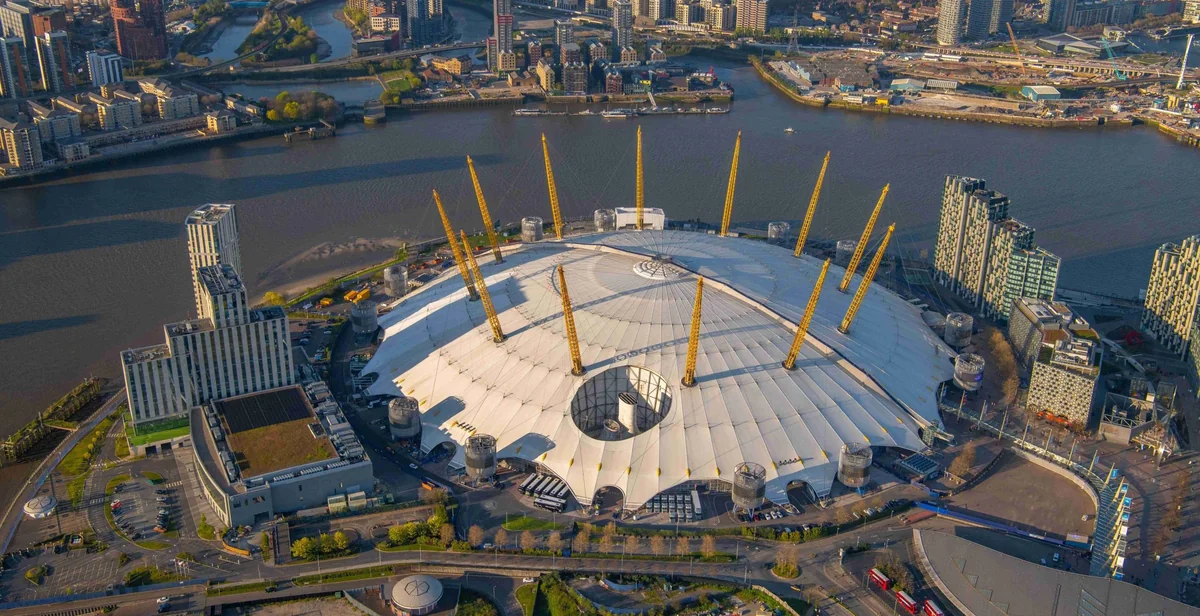The aerial view shows the London 2012 Olympics' iconic White Cube, a large white dome with yellow accents, surrounded by a circular concave and a river. The cityscape features modern buildings and a bridge, with the London 2012 Olympics as the central focus. The image captures the city's blend of modernity and tradition, with the White Cube symbolizing the city's spirit.