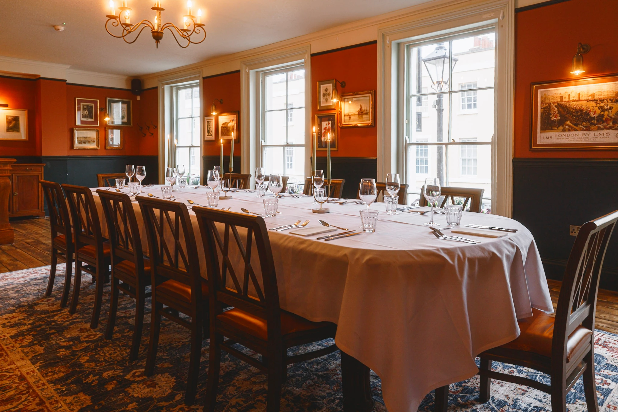 The image shows a luxurious dining room with a long, white-clothed table set for six, surrounded by wooden chairs. The room features red walls, framed pictures, and chandeliers, with large windows allowing natural light. The floor is covered with a patterned rug, and the room exudes elegance and sophistication.