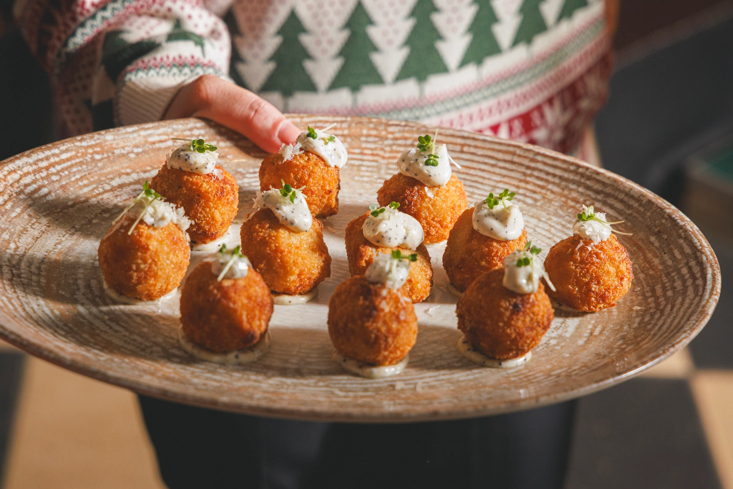 A hand in a white and green sweater holds a woven plate with nine golden-brown, fried dough balls topped with white sauce and garnished with green herbs. The plate is set against a wooden floor, creating a warm and inviting atmosphere.