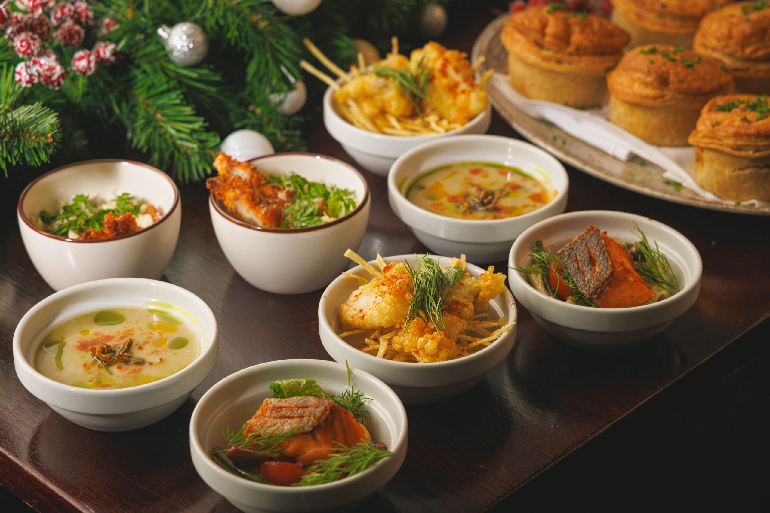 A festive table setting features a variety of dishes, including soup, fish, quiche, and tartar sauce, with a tray of pastries and a Christmas tree in the background. The arrangement is harmonious and inviting, with the soup and tartar sauce as focal points.
