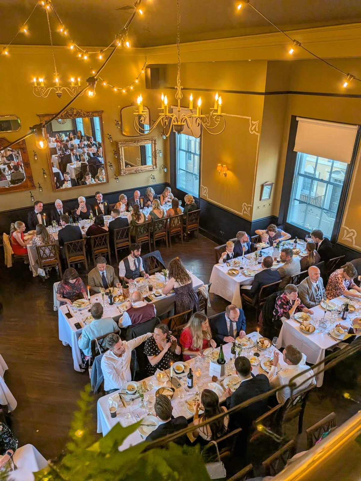 A high-angle image captures a bustling banquet hall with 20 tables draped in white tablecloths, each adorned with plates and glasses. The room is illuminated by chandeliers and warm lighting, with a large mirror reflecting the room's ambiance. The high vantage point offers a comprehensive view of the event, capturing the lively atmosphere and the anticipation of the guests.