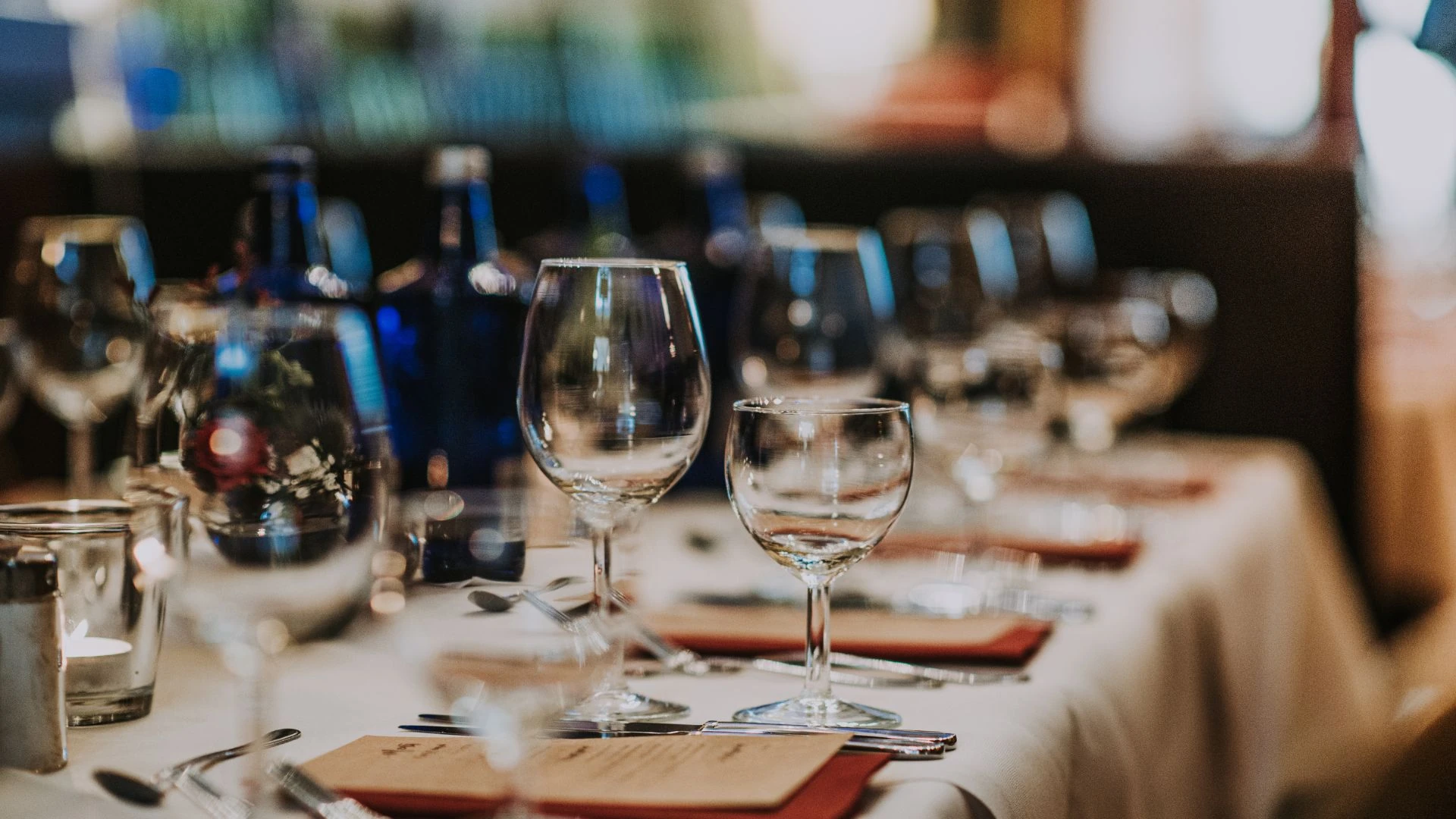 The image shows a beautifully set table for a formal dinner, with a white tablecloth, red napkins, and clear glasses. Blue bottles with flowers and silverware are arranged neatly. The blurred background suggests a restaurant or bar setting, creating anticipation for a sophisticated dining experience.