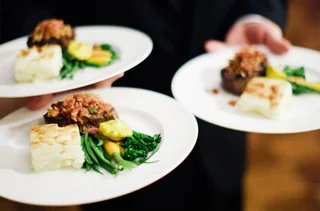 In the image, two hands hold white plates with food, showcasing a variety of dishes. The plates have white plates with a variety of food, including meat, vegetables, and a green salad. The hands are in the foreground, with the plates in the background. The background is blurred, emphasizing the focus on the food. The image captures a moment of anticipation and anticipation.