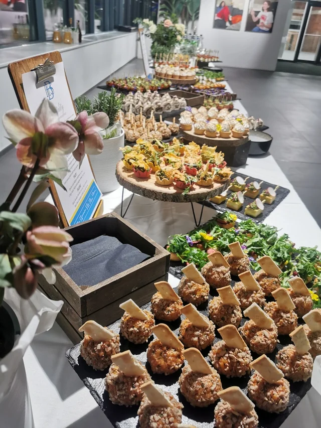 The image features a well-organized buffet table with a white tablecloth, featuring a variety of food items in wooden boxes and trays. The food includes fried balls, salads, and sandwiches. A blue menu is displayed on the table, and a large flower arrangement adds a touch of nature. The background shows a room with a window and a large window, creating a spacious atmosphere. The image captures a moment of anticipation before the feast begins.