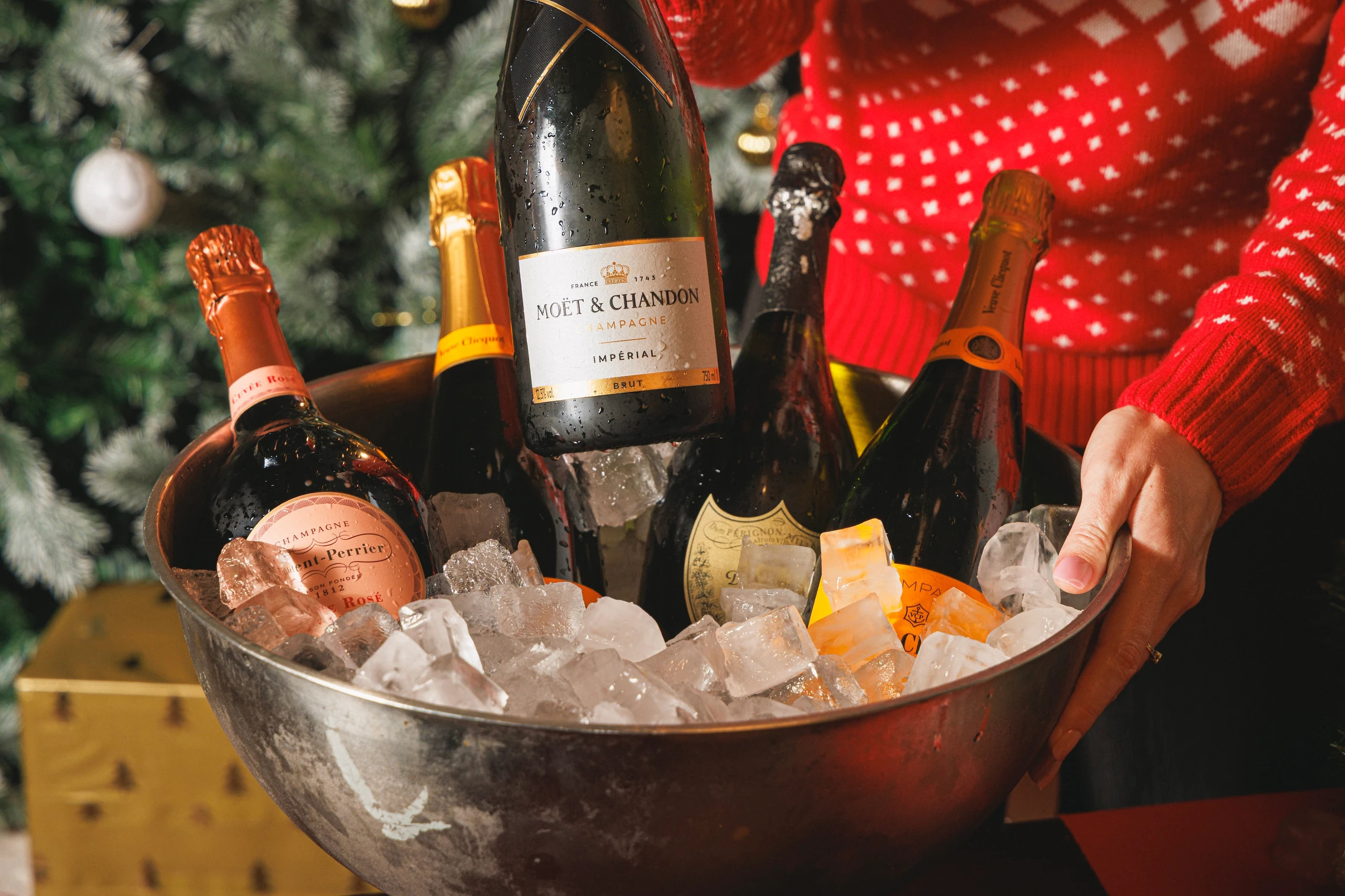 A person in a red sweater holds a silver bowl filled with ice cubes and a Christmas tree in the background. A bottle of Chateau Marnier "Chateau Marnier" wine is in the bowl, with a bottle of champagne nearby. The scene suggests a festive celebration, possibly a holiday gathering.