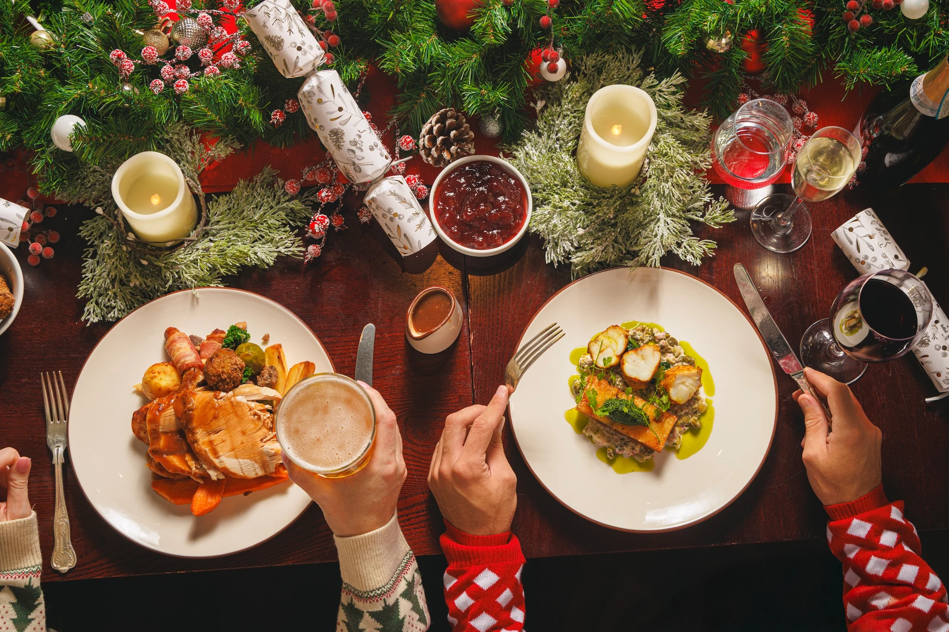 A festive table setting features two plates of food, a glass of red wine, a glass of beer, and a plate of potatoes. Two hands are holding the plates, with one wearing a red and white striped shirt. The table is adorned with Christmas decorations, including pine cones, a wreath, and a garland. The scene is set against a backdrop of a Christmas tree and a red ribbon.