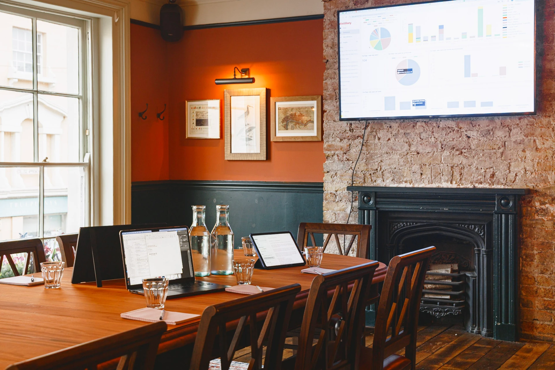 The image shows a cozy restaurant with warm orange walls, a fireplace, and a large window. A wooden table with wine glasses and menus is in the center, surrounded by wooden chairs. A mounted television displays a graph, and framed pictures adorn the walls. The room is well-lit and inviting, with a sense of organization and style.