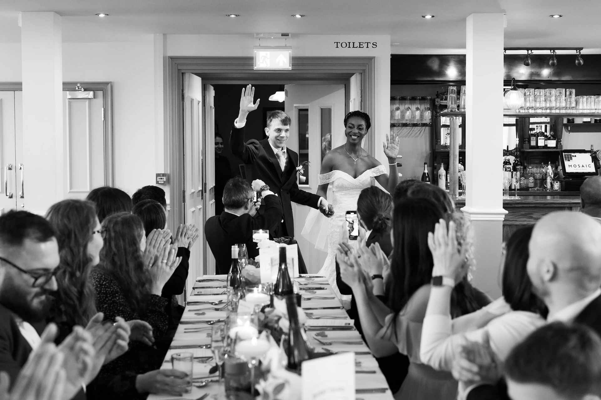 A black and white photo captures a joyous wedding reception with a bride in a white dress and groom in a black suit. They are at a long table with elegant tablecloths and wine glasses, and the bride is clapping, while the groom is smiling. The background shows guests in formal attire, and the photo is taken from a low angle, emphasizing the couple's happiness. The image is rich in detail and captures the essence of a special occasion.