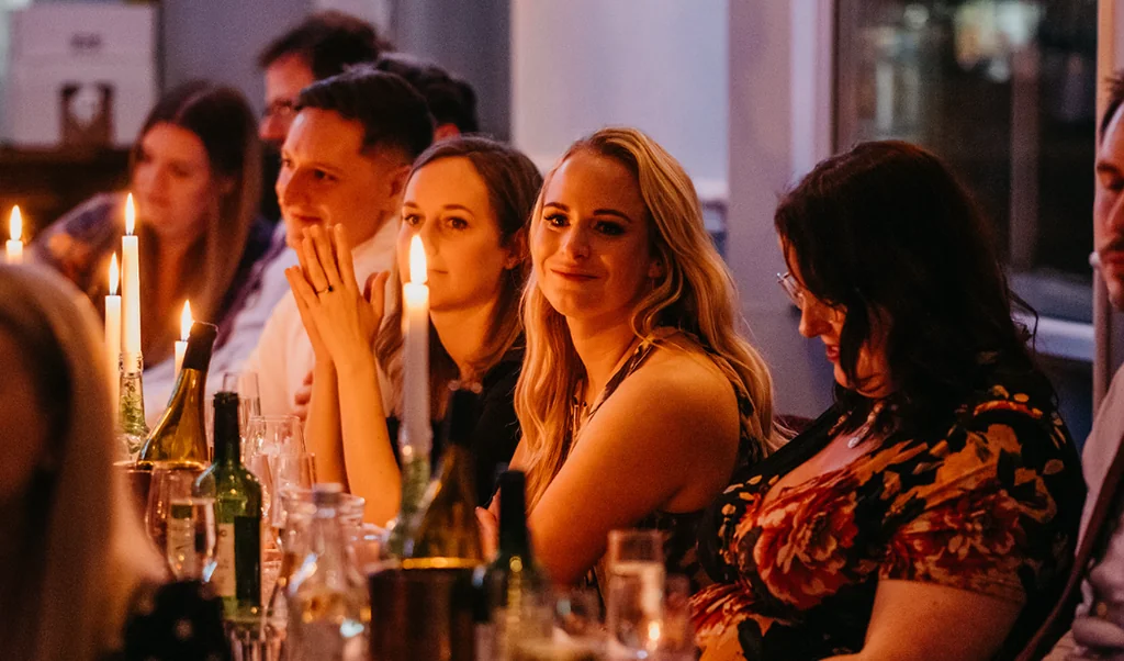 In a dimly lit room, a group of people gather around a long table adorned with candles and wine bottles. A woman in a floral top and a man in a suit are present, with the woman's hand on her chin and the man's hand on his cheek. The table is filled with various objects, including wine bottles, glasses, and a bottle of wine. The scene is set against a wall with a picture and a window, creating a warm and intimate atmosphere.