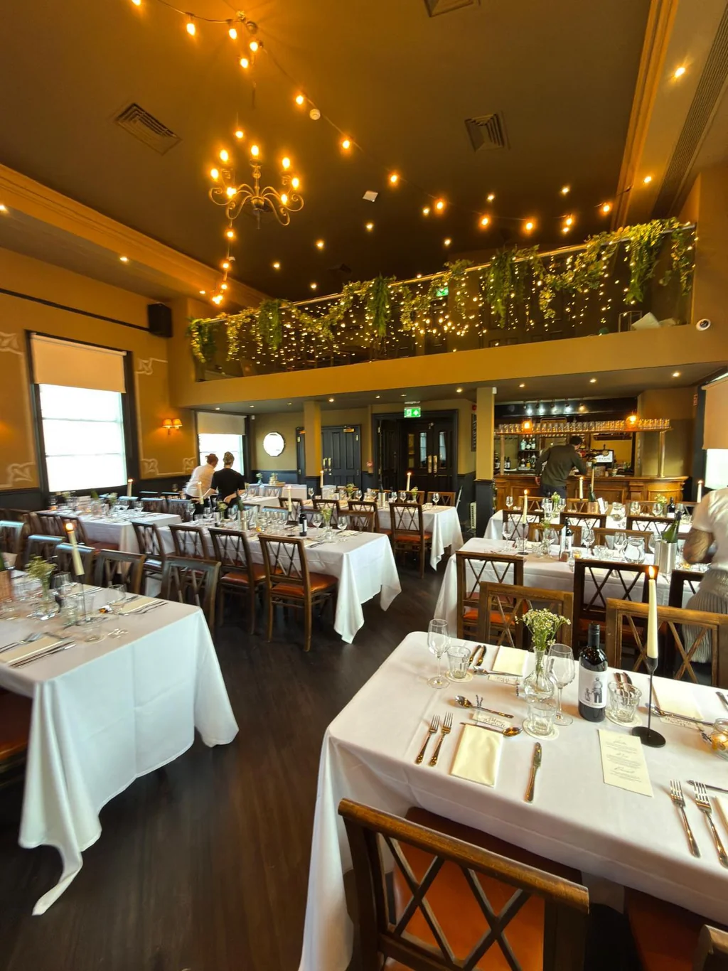 The image shows a spacious, well-lit restaurant with a high ceiling adorned with greenery and string lights. Tables with white tablecloths and silverware are arranged in rows, with wine glasses and silverware on each table. A chandelier hangs from the ceiling, and a bar is visible in the background. The restaurant is empty, creating a serene atmosphere.