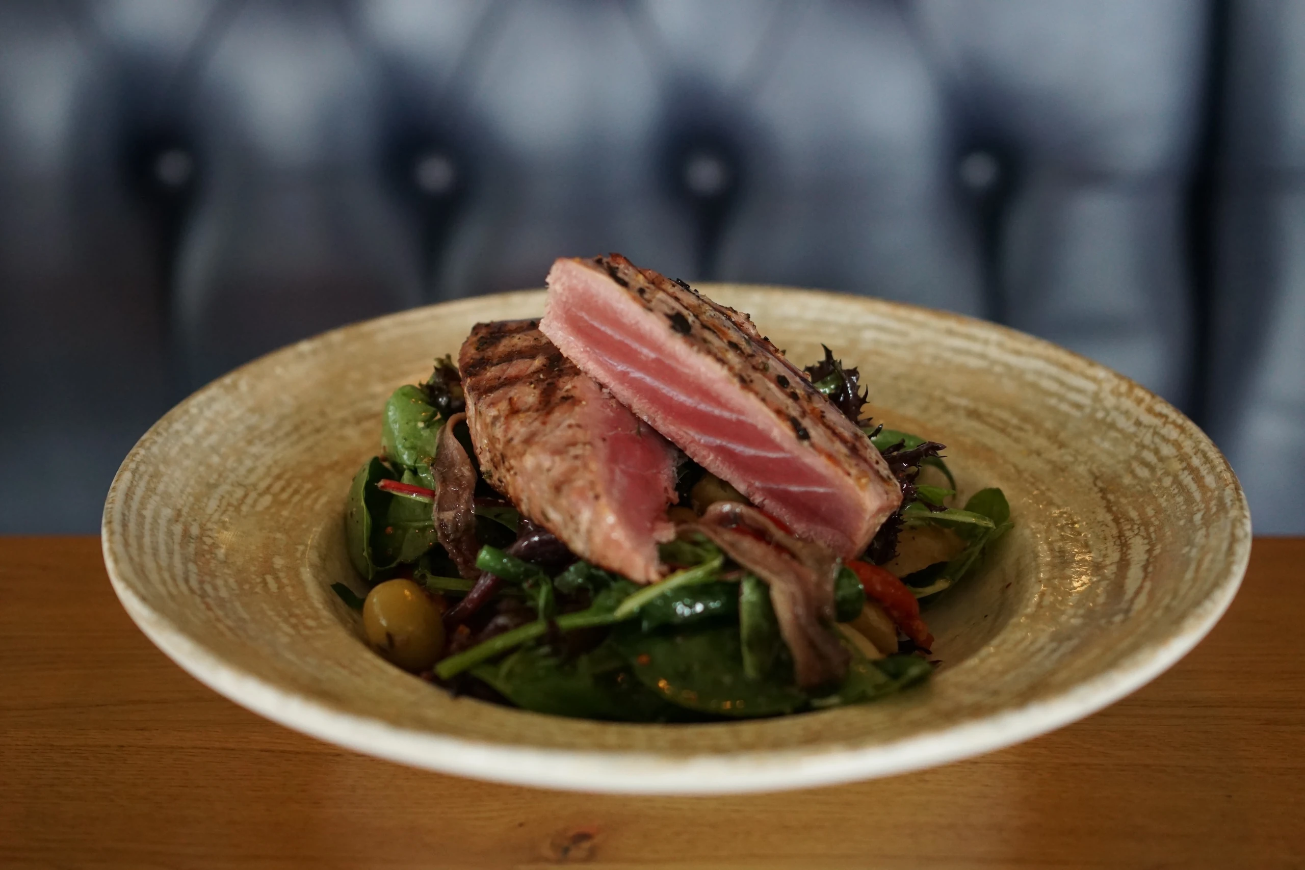 A white plate with a grilled steak, garnished with herbs and a red sauce, is the centerpiece of a colorful salad. The steak is on the left side, while the salad is on the right. The plate is on a wooden table, with a blue leather booth in the background. The scene is visually appealing and suggests a restaurant setting.