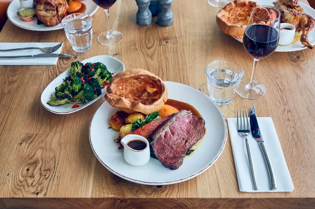 A wooden table features a large, dark brown steak with a rich sauce, surrounded by a variety of dishes including roasted potatoes, green vegetables, and a roasted meat. Two glasses of red wine are nearby, and a white plate with a silver fork and knife is on the left side. The arrangement creates a balanced and appetizing scene.