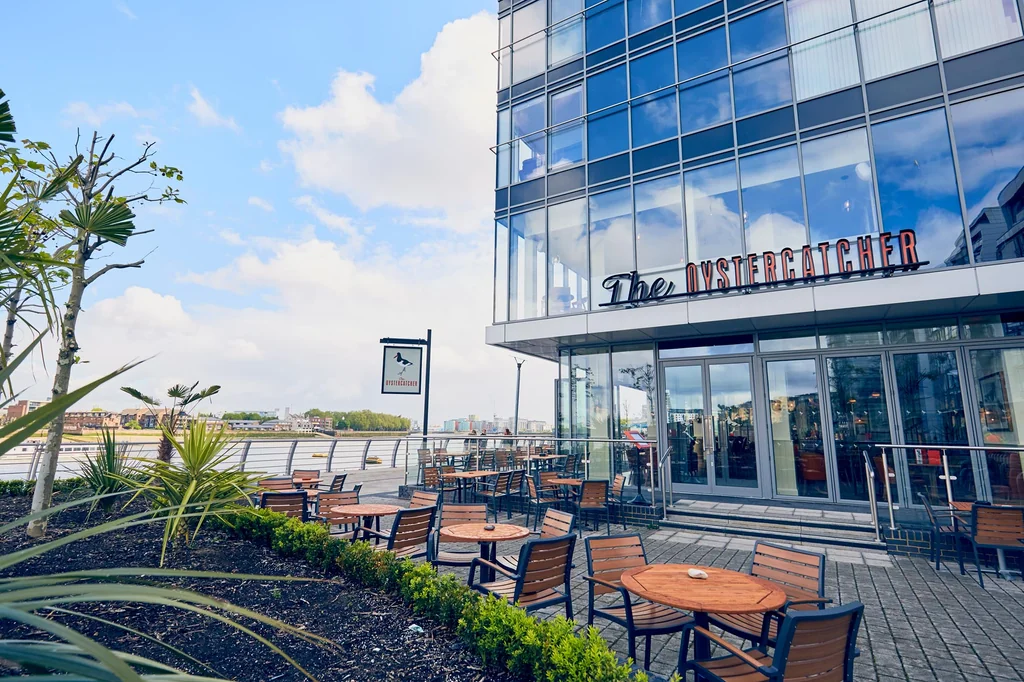 The image features a modern glass and steel building with a "Mallory" sign, overlooking a waterfront. The building is surrounded by a patio with wooden tables and chairs, surrounded by plants and a fence. The sky is a clear blue with scattered clouds, and the city skyline is visible in the distance. The scene is serene and inviting, with a harmonious blend of urban and natural elements.