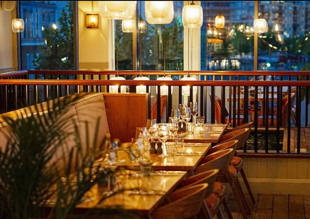 A serene restaurant interior features a wooden table with six chairs, set for a meal. The table is adorned with silverware and a vase of greenery. The room is illuminated by hanging lights and a wooden railing. The view outside shows a cityscape, creating a tranquil atmosphere.