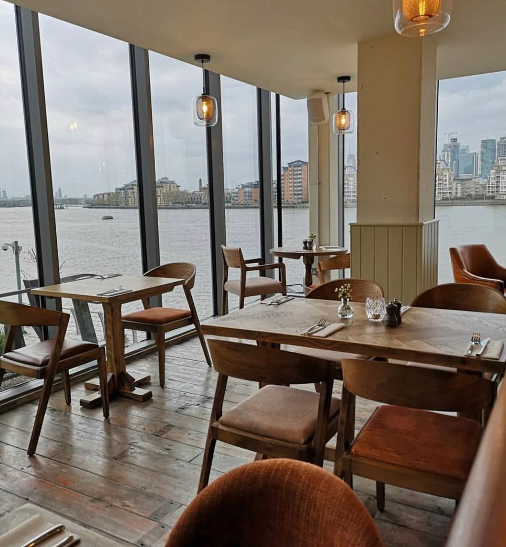 The image shows a modern restaurant with a large window overlooking a body of water, offering a panoramic view of the city skyline. The restaurant features wooden tables and chairs, with white tablecloths and silverware. The room is well-lit, and the atmosphere is serene and inviting.