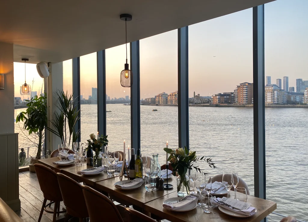 The image shows a modern restaurant with a large window overlooking a city skyline, featuring a wooden dining table with white plates, silverware, and wine glasses. The table is set with white napkins, silverware, and a vase of flowers. The restaurant has a modern design with a wooden floor and a large window. The city skyline is visible through the window, and the restaurant is illuminated by a hanging light fixture. The scene is serene and inviting, with a harmonious blend of modern and natural elements.