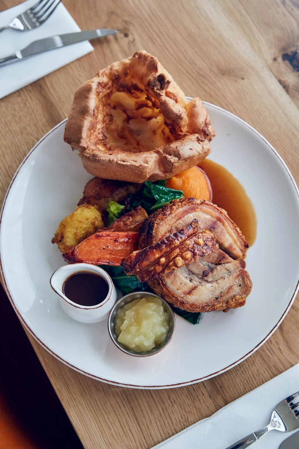 A wooden table features a white plate with a variety of food items, including a roast, mashed potatoes, carrots, and a side of sauce. Utensils are arranged around the plate, and a napkin is nearby. The scene is visually appealing and inviting.