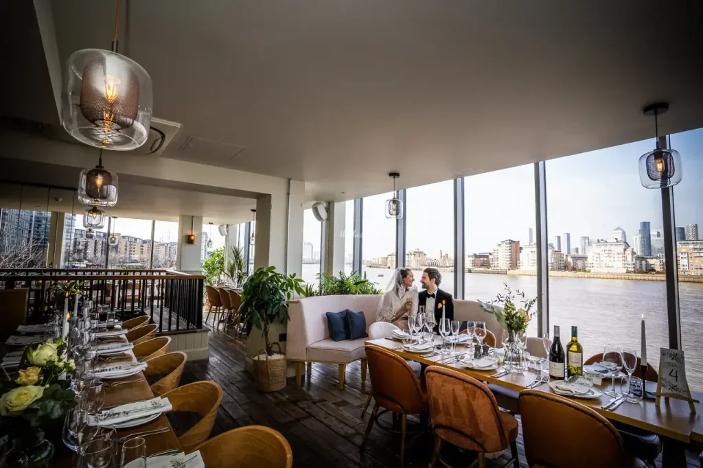 A modern restaurant with a river view is illuminated by hanging lights and a large window. Two couples are seated at a table with elegant tablecloths and wine glasses, while a white sofa and wooden chairs provide seating. The restaurant is well-lit and has a modern aesthetic, with a river view and a city skyline in the background.