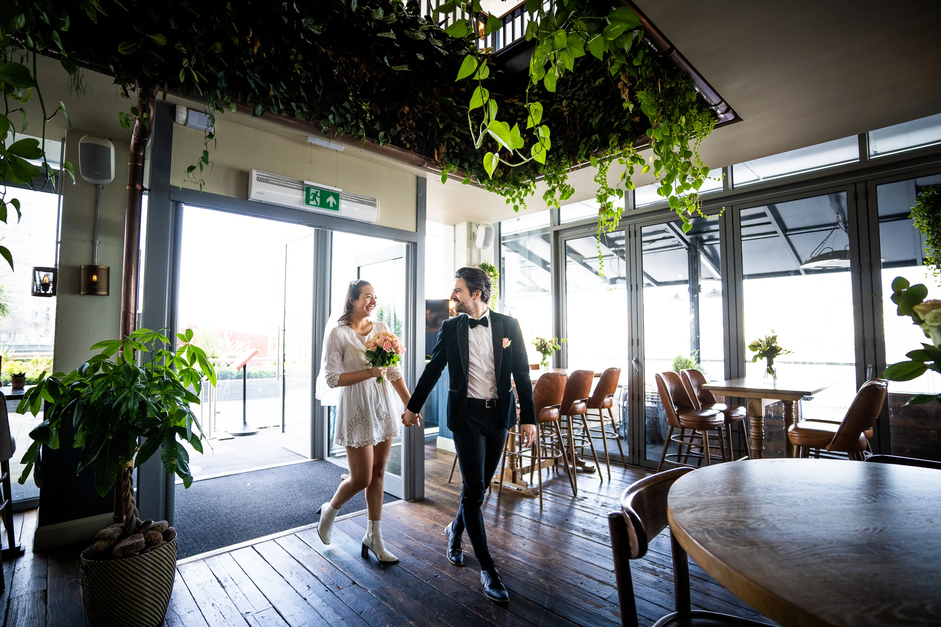A couple in formal attire walks hand in hand through a modern, glass-walled restaurant with a high ceiling and large windows. The man wears a black tuxedo and the woman a white dress. The restaurant has a bar area with wooden tables and chairs, and potted plants add a touch of nature. The scene is bathed in natural light, creating a warm and inviting atmosphere.