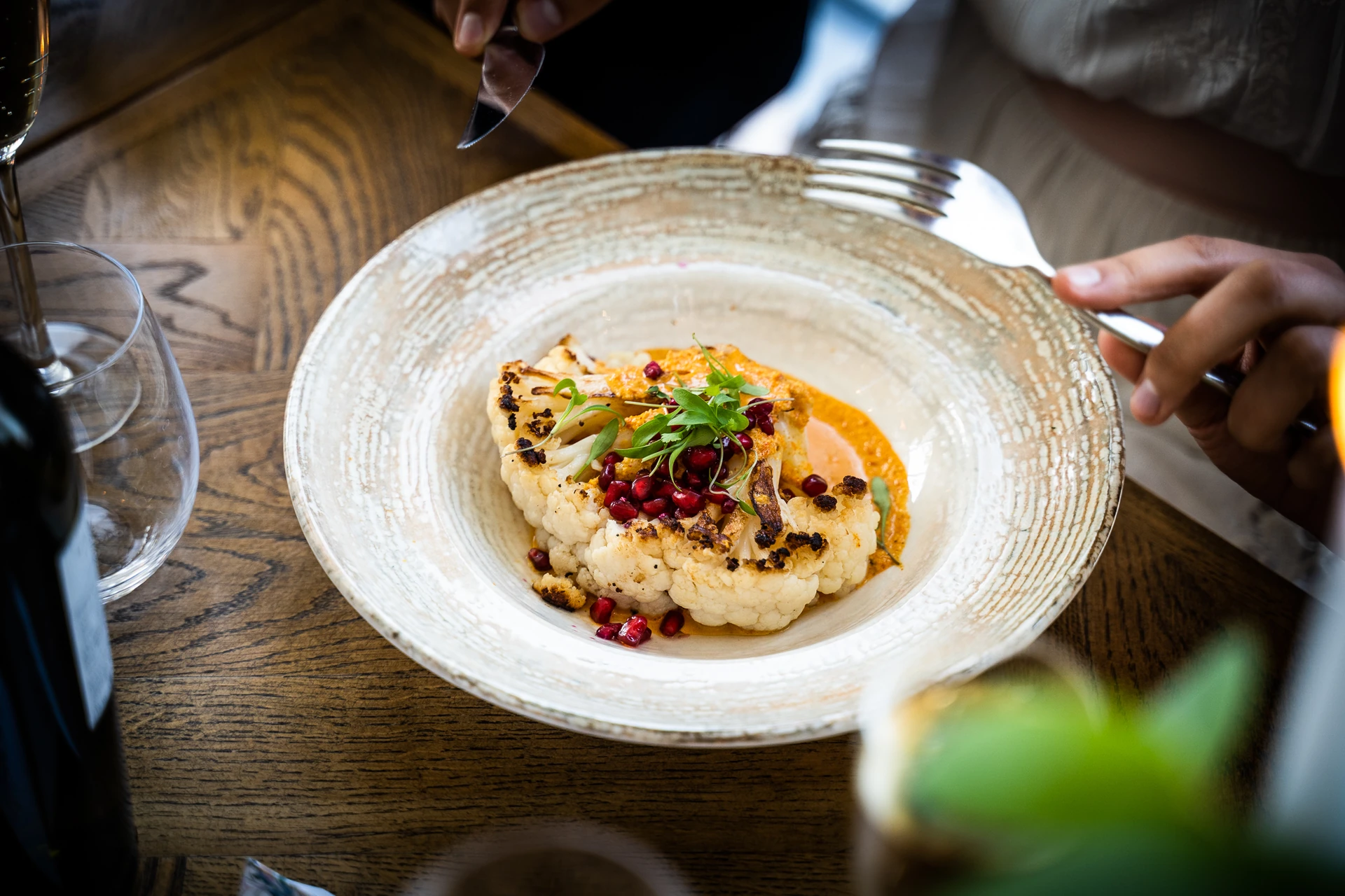A white plate with a golden rim holds a dish of white fish with red and green garnishes, accompanied by a wine glass and a fork. The scene is set on a wooden table with a plant in the background, creating a warm and inviting atmosphere.