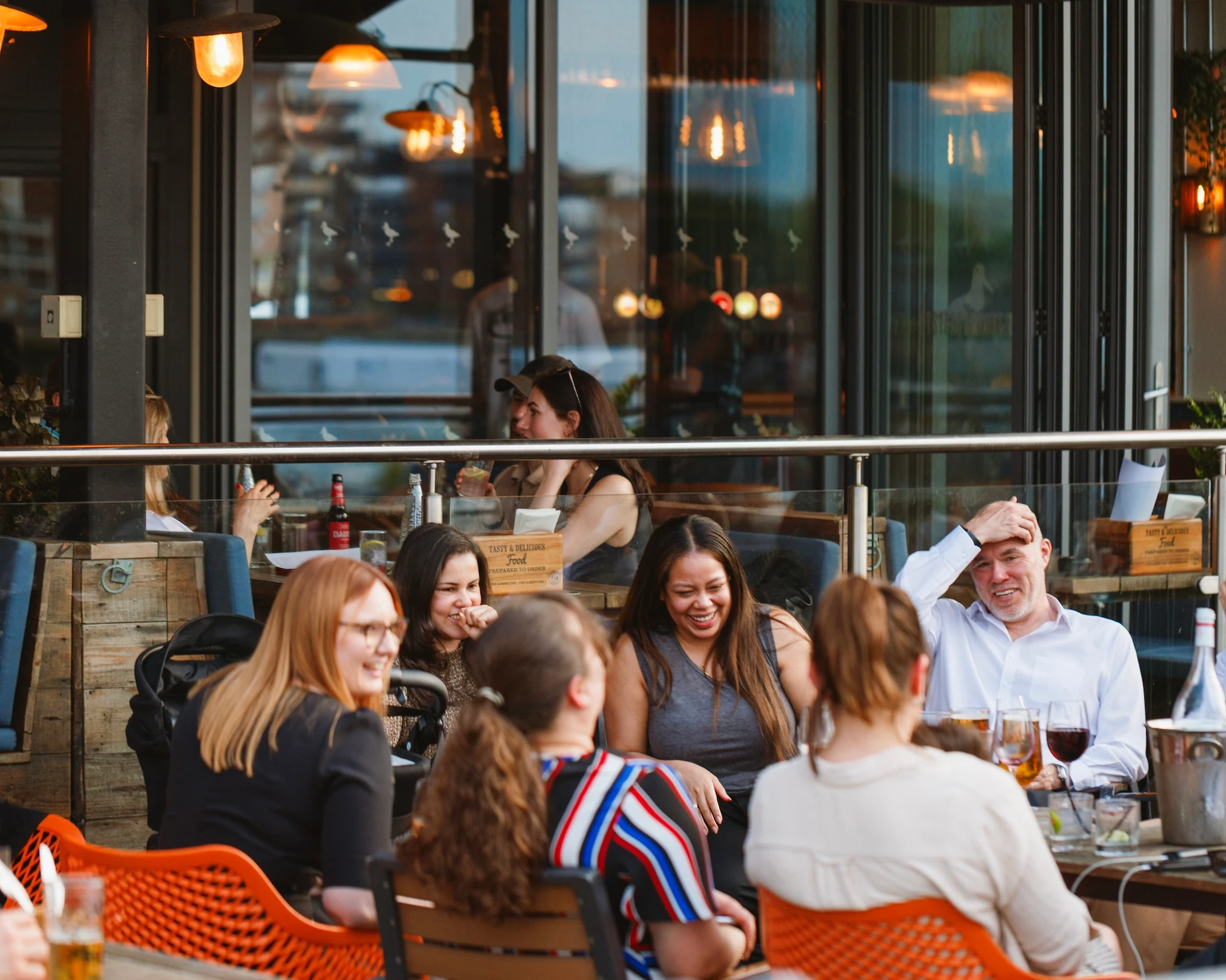 A lively outdoor restaurant scene features a group of people enjoying a meal and conversation, with a woman in a striped shirt and a man in a white shirt. The restaurant has a large glass window and a metal bar, and the atmosphere is warm and inviting.