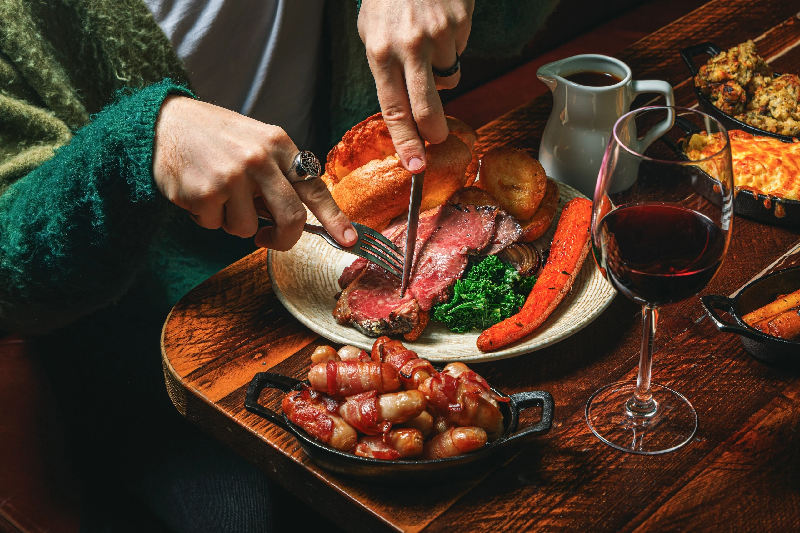 A person in a green sweater is cutting meat on a white plate with bacon and green vegetables, using a knife and fork. A white pitcher and a glass of red wine are on the table, with a black serving tray and a white plate of bread nearby. The scene is set against a dark background, emphasizing the meal and the person's focus.