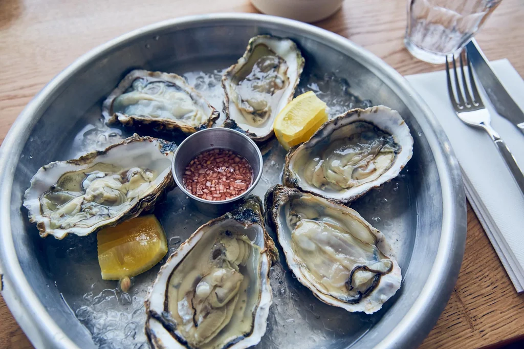 A silver tray on a wooden table holds six oysters, each with a white shell and green crust, surrounded by lemon slices and red pepper flakes. A glass of water and a fork are nearby, creating a casual dining atmosphere.