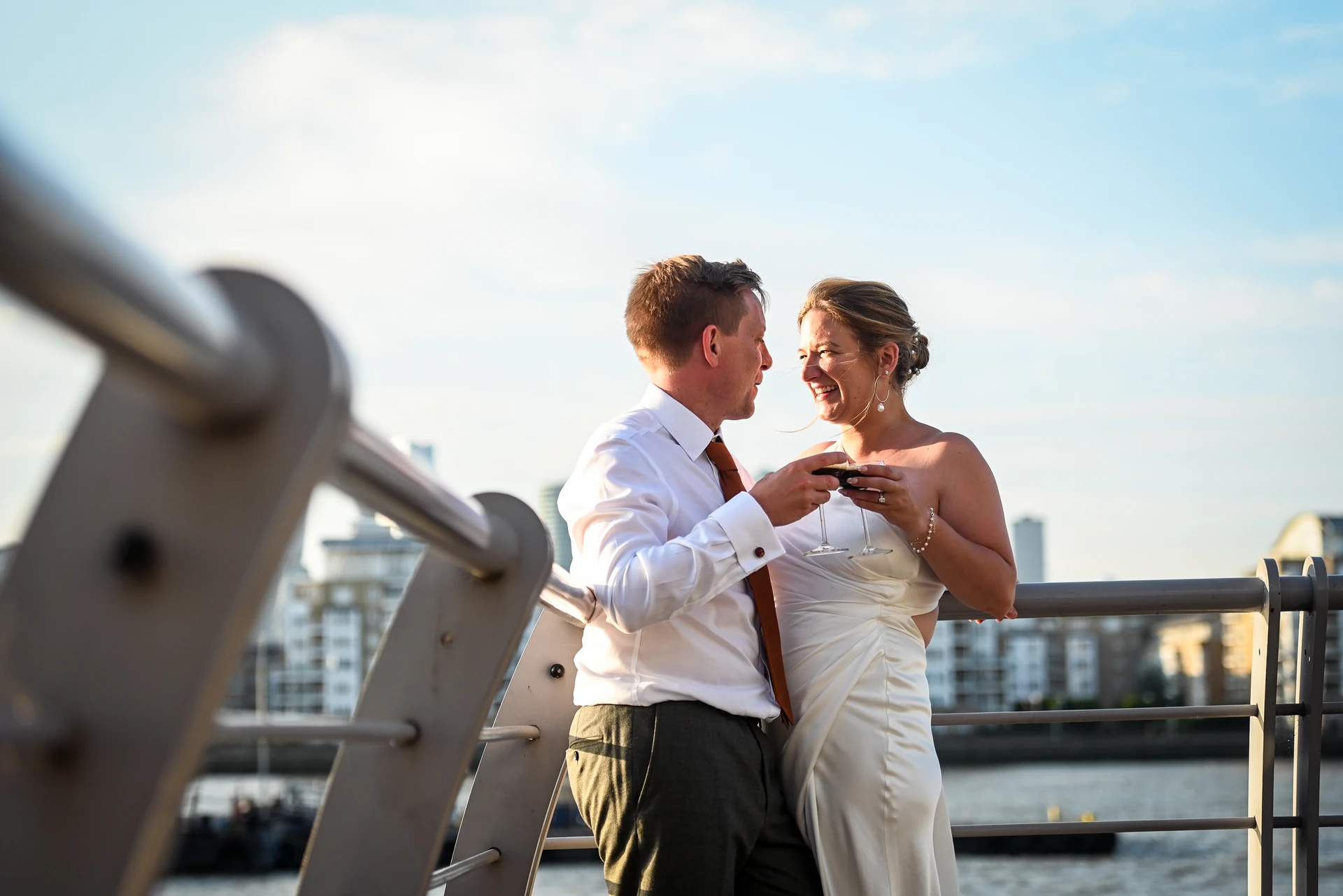 A couple in a white wedding dress and red tie, with the man holding a cell phone, stand on a metal railing overlooking a body of water. The woman wears a white dress with a silver necklace and a bracelet. The background features a cityscape with white buildings and a blue sky with clouds.