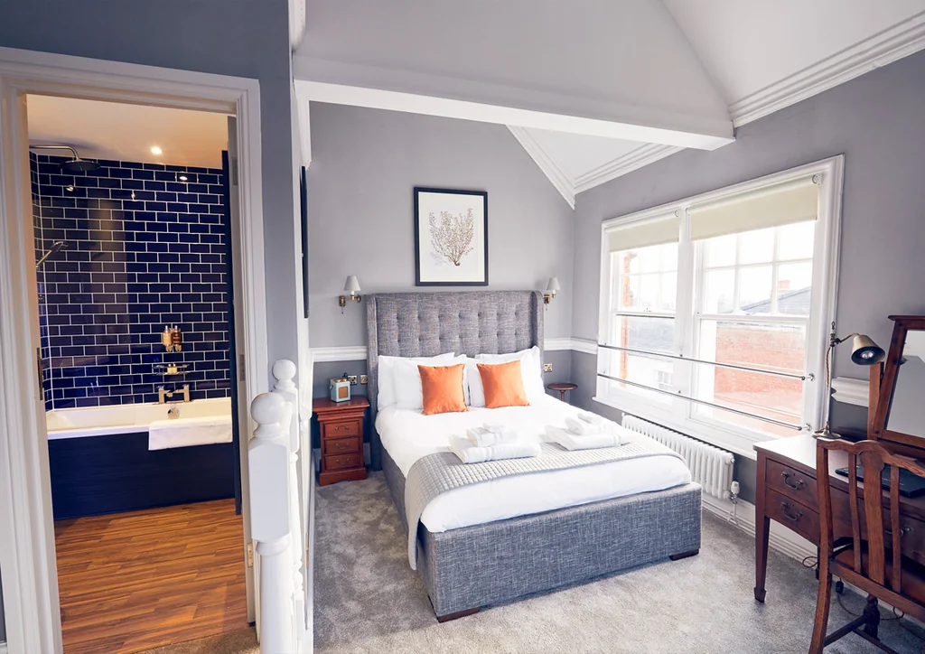 The image shows a modern bedroom with a large gray bed, white linens, and orange and white pillows. A wooden desk and chair are to the right, with a white radiator and lamp on the left. A blue and white painting hangs above the bed, and a window with white curtains allows natural light. The room has a minimalist aesthetic with a white radiator, white radiator, and white radiator. The floor is gray carpet, and the walls are gray. The room is well-lit and has a modern design.