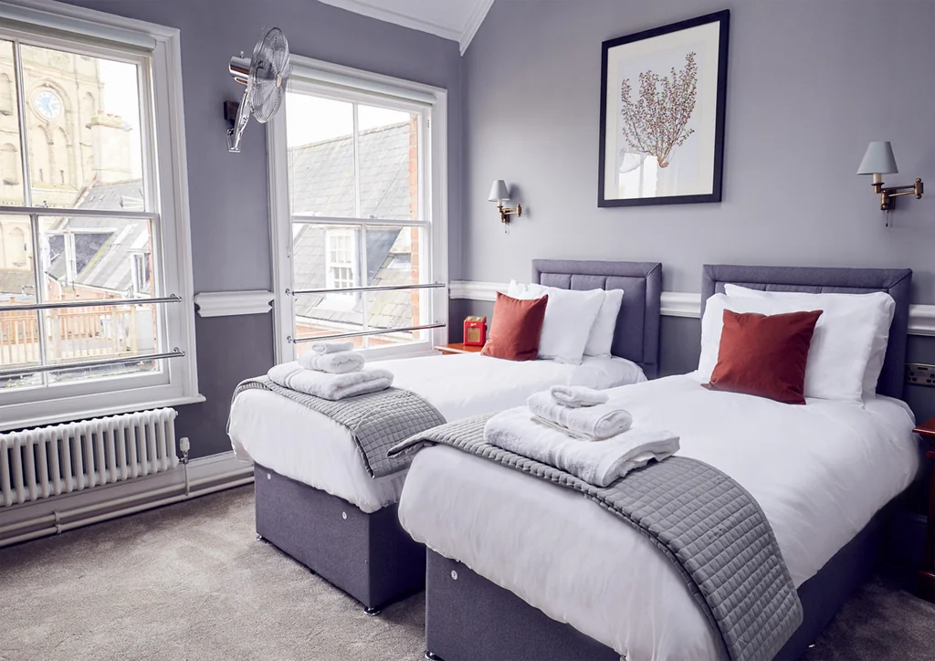 The image shows a serene bedroom with two twin beds, each adorned with white linens and red pillows. The beds are positioned next to a window, offering a view of a building outside. The room features a white radiator, a framed picture of a tree, and a lamp with a glass shade. The color scheme is neutral, with white, gray, and red accents. The room is well-lit, creating a tranquil atmosphere.