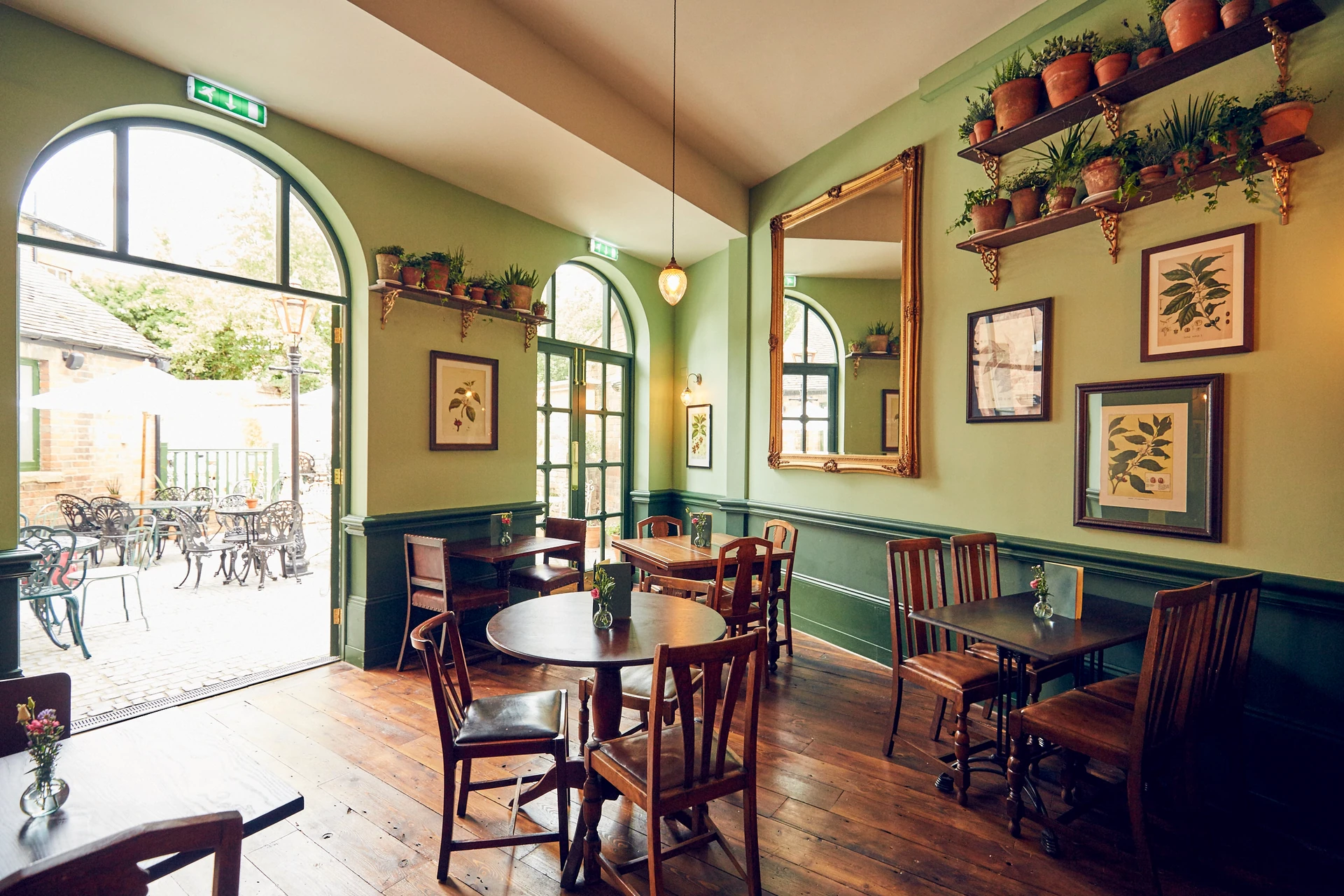 The image shows a cozy, rustic pub with green walls and wooden floors, featuring wooden tables and chairs, potted plants, and a large arched window. The pub is located on a patio with a garden, and the entrance is marked with a green sign. The scene is warm and inviting, with a harmonious blend of nature and architecture.