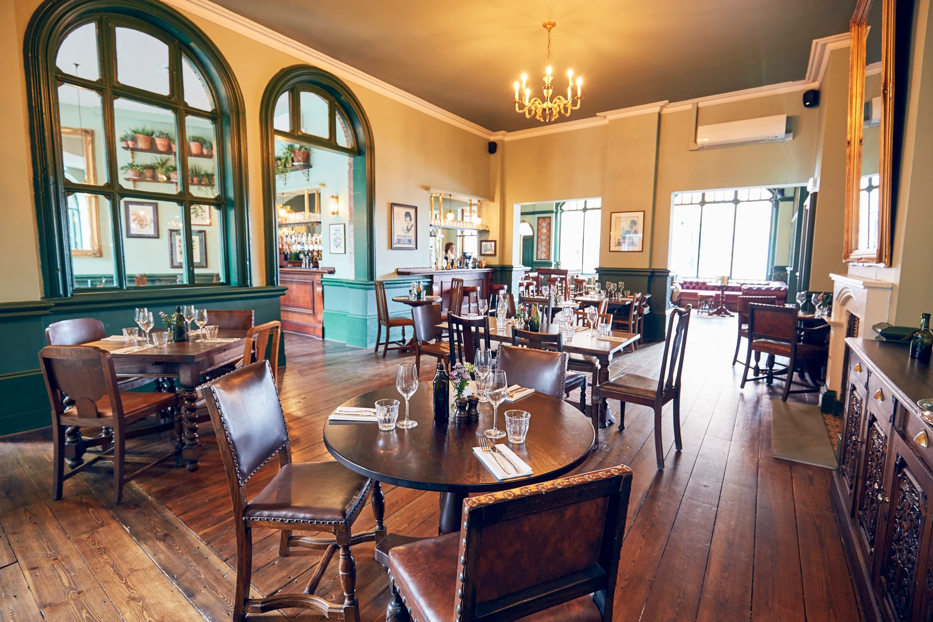 The image shows a luxurious dining room with a warm, inviting atmosphere. The wooden floor and chairs contrast with the green walls, while the large windows offer a view of the outside. The room is meticulously set with white tablecloths, silverware, and wine glasses, and features a chandelier and mirror. The layout is harmonious, with the dining area on the left and the bar on the right. The room exudes elegance and sophistication, making it a perfect place for social gatherings.