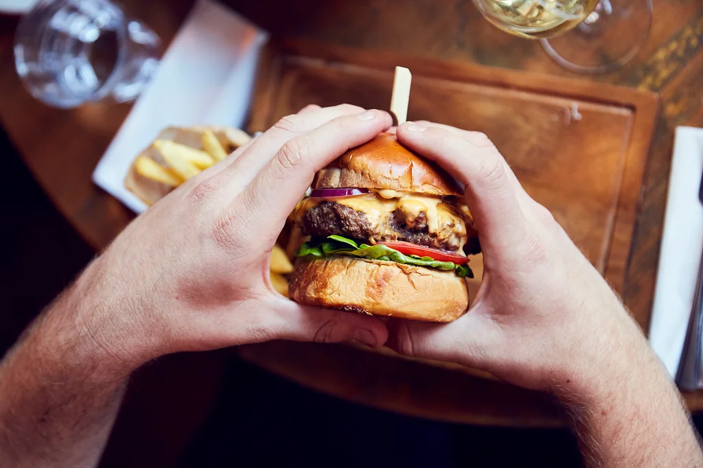 A person is seated at a wooden table, holding a large, well-made hamburger with various toppings. The table has a white plate, a glass of wine, and a tray of fries. The person's hands are holding the hamburger, and the background is blurred, emphasizing the hamburger and table setting.