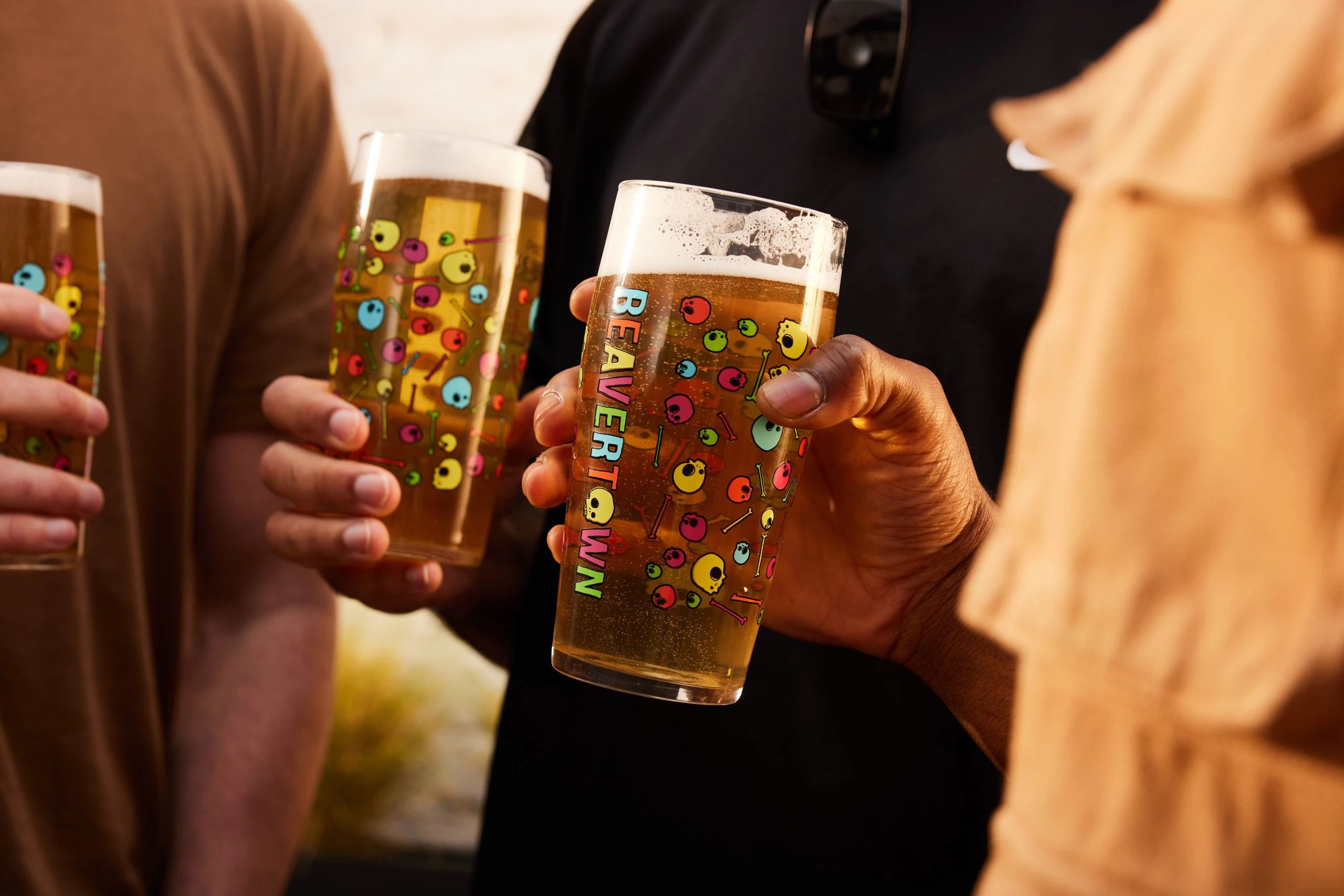 Three individuals in a relaxed setting, each holding a half-filled glass of beer, are dressed casually in a tan shirt and a black shirt. The beer is golden yellow, with a frothy head and colorful designs. The background is blurred, emphasizing the beer and the individuals. The image captures a casual, social gathering with a sense of camaraderie.