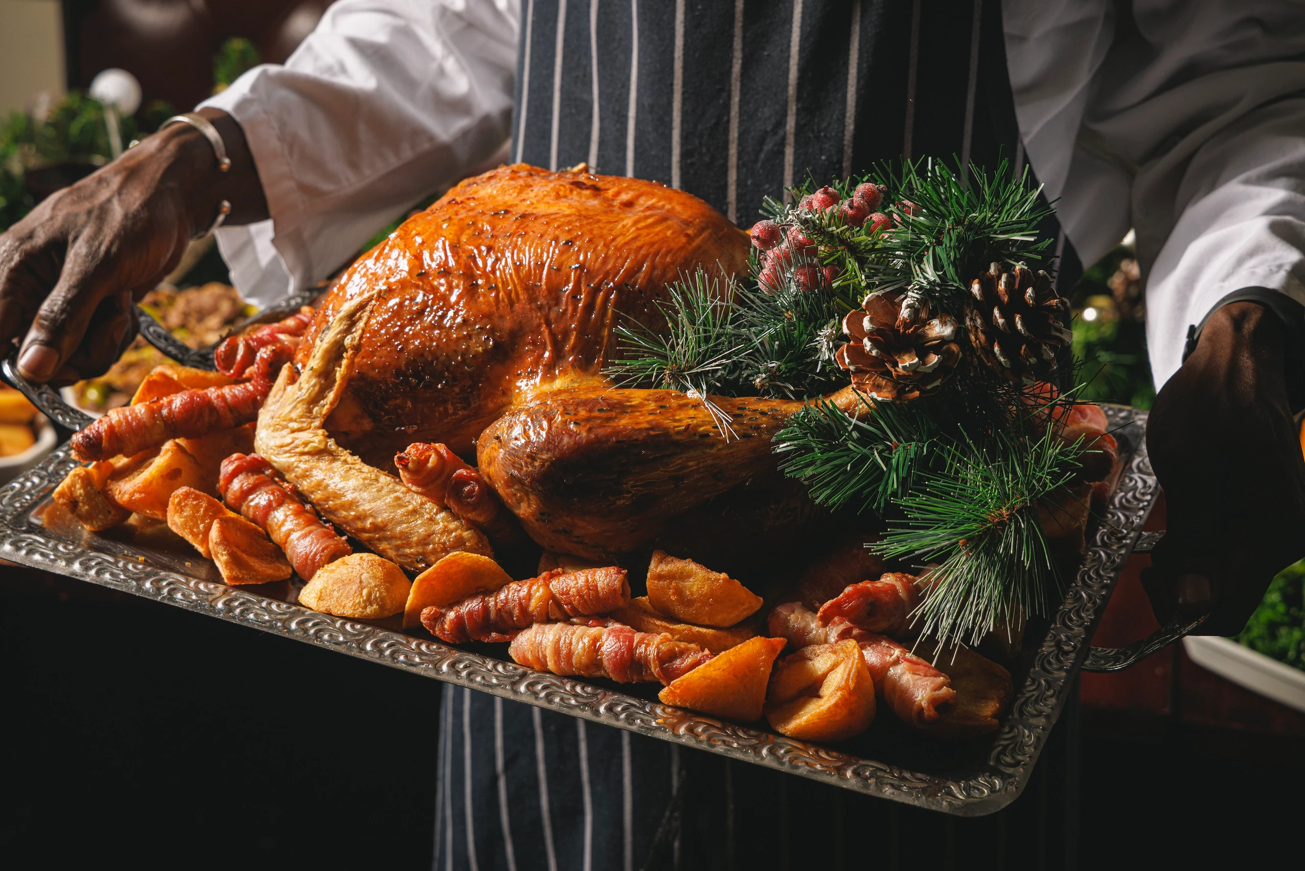A person in a white apron and black striped shirt holds a large, golden-brown roasted turkey on a silver tray, surrounded by carrots, bread, and pinecones. The turkey is the centerpiece, with the person's hand holding the tray. The background features a festive setting with a Christmas tree and pinecones. The image captures a moment of anticipation before a festive feast.
