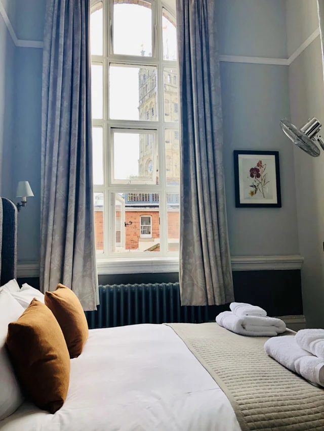 The image features a serene bedroom with a large window, white curtains, and a radiator. The bed is neatly made with white linens and two brown pillows. A framed picture hangs above the window, and a lamp is on the right side. The room is bathed in soft light, creating a tranquil atmosphere.